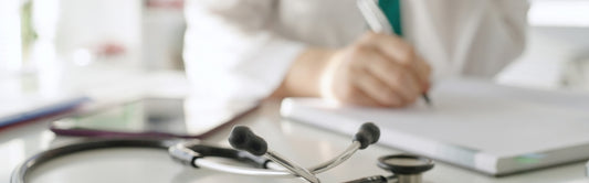 Ein Stethoskop liegt auf einem Schreibtisch im Vordergrund, während eine Person in einem weißen Kittel im Hintergrund auf einem Notizblock schreibt.