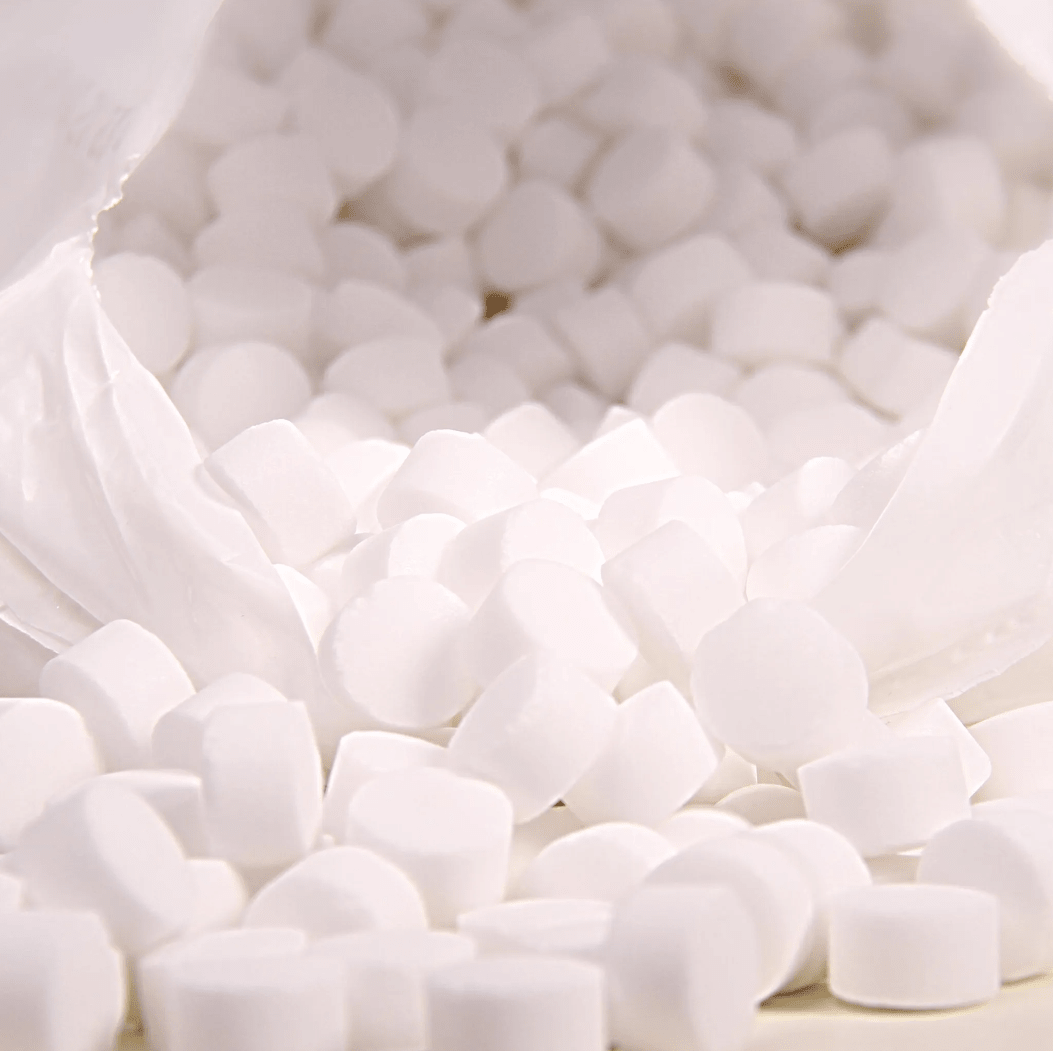 Un primer plano muestra una bolsa de plástico blanca con la inscripción «K+S Minerals and Agriculture GmbH», que contiene pastillas de sal de evaporación Regenit conformes a la norma EN 973, tipo | saco (25 kg), las cuales caen sobre una superficie. Las pastillas, pequeñas y redondas, tienen una forma y un tamaño uniformes y son ideales para su uso en sistemas de descalcificación de agua.