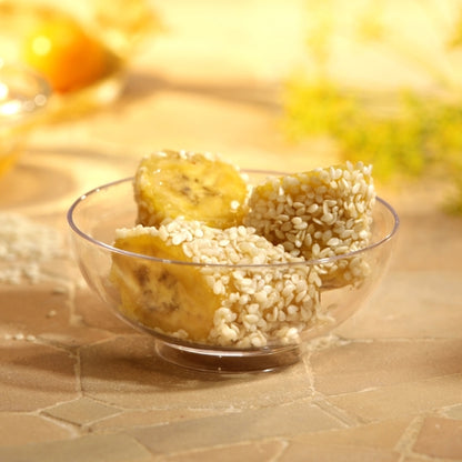 Three pieces of banana with white sesame are served in a Starpak 50 finger food bowl (PS clear), placed on a light tile surface with a gently blurred background – ideal for events.