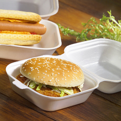 Una hamburguesa en un panecillo de sésamo con lechuga y hamburguesa, servida en una caja para hamburguesas Starpak 100 con tapa abatible, XPS, 7,5 x 12 x 12 cm blanca - ideal para catering, con perritos calientes cerca y hojas de lechuga en una superficie de madera.