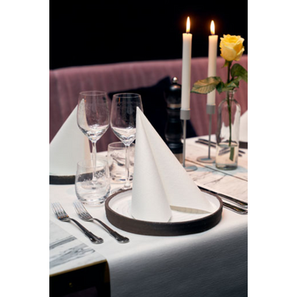 A formal table arrangement includes neatly folded Dunilin® napkins 40 x 40 cm from Duni GmbH on each plate, complemented by wine glasses, a water glass, cutlery, burning candles, and a single yellow rose in a vase. A light pink fabric serves as the background.