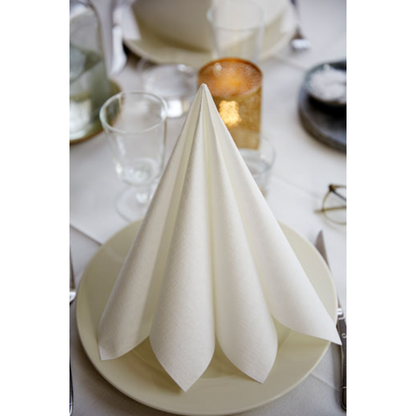 A neatly folded Dunilin® napkin 40 x 40 cm from Duni GmbH stands upright on a white plate, surrounded by glasses and cutlery on a set table. A burning candle and a few glasses enhance the ambiance with sustainable table decoration in the background.