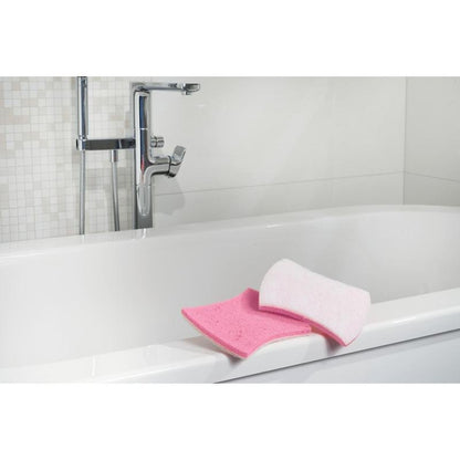 Shown is a white bathtub with chrome fittings. On the edge are a pink bathroom sponge and a Scotch-Brite® Bath XXL cleaning sponge for the bathroom from 3M Deutschland GmbH. The background shows white tiles with a mosaic pattern.