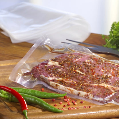 Ein Starpak 100 Siegelrandbeutel, PA/PE transparent von Starpak hält mariniertes rohes Fleisch auf einem Holzbrett mit grünen und roten Chilischoten, Zangen und Blattgrün - ideal für Ihren Kammervakuumierer.