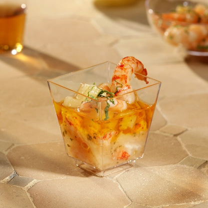 A Starpak 50 finger food bowl (PS clear) from Starpak, filled with shrimp and seafood cocktail and garnished with herbs, stands on a beige, textured surface. More cups are blurred in the background – perfect for stylish events.