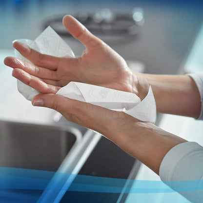 A person dries their hands over a kitchen sink with Scott® Essential™ paper towels - roll / white (6 rolls) from Kimberly-Clark GmbH. Blue graphic lines accentuate this practical hygiene solution in front of a blurred countertop and a faucet.
