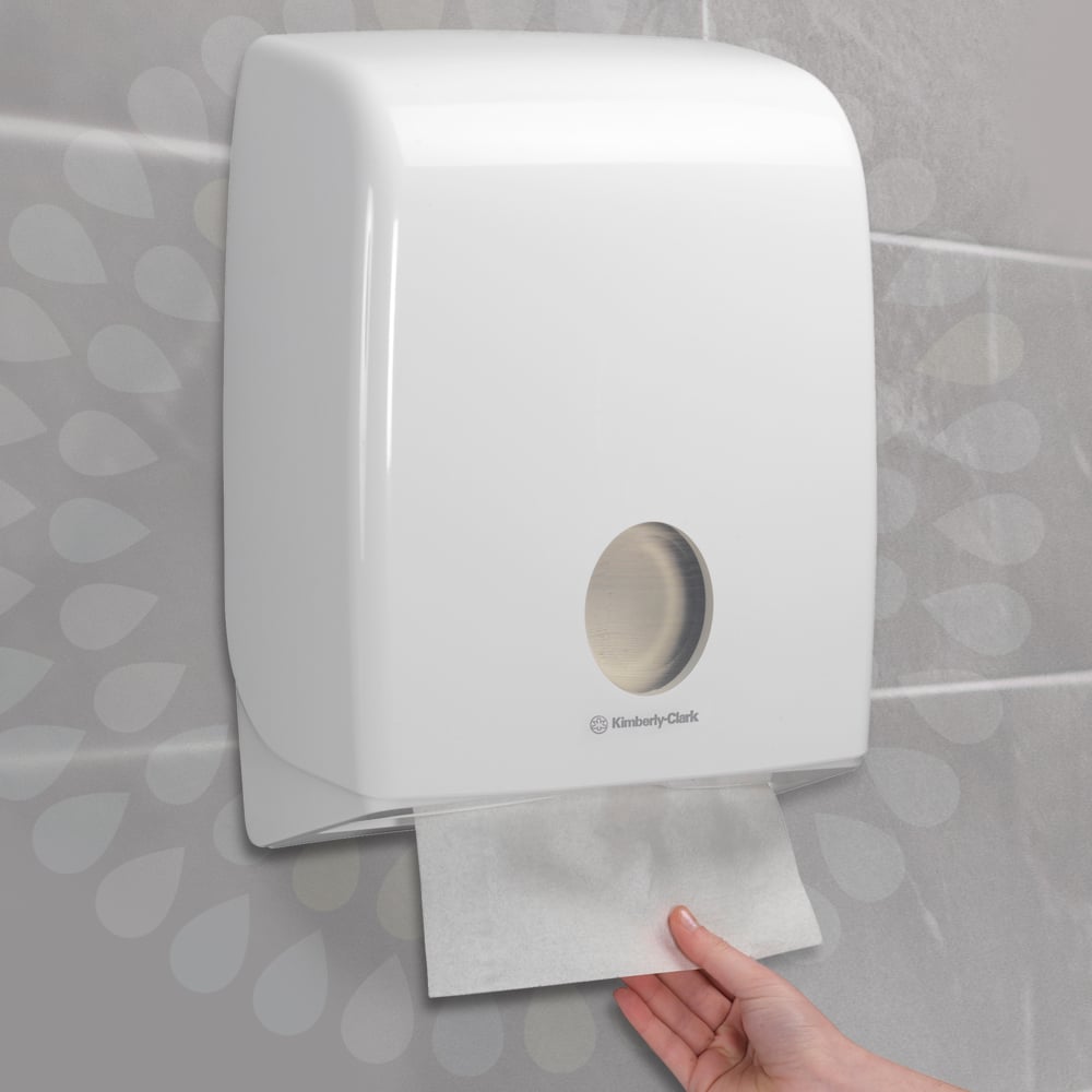 A hand pulls a Kleenex® Ultra™ Hand Towel from a wall-mounted white dispenser by Kimberly-Clark GmbH, installed on a tiled wall (Interfold/White/Large - 30 clips x 94 sheets | box, 30 packs).