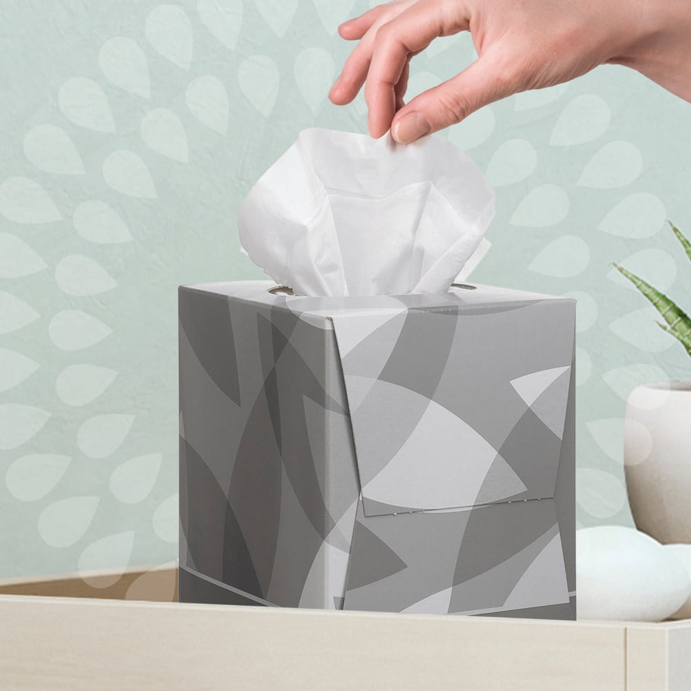 Eine Hand zieht ein Kleenex® Kosmetiktücher - Würfel-Box / Weiß von Kimberly-Clark GmbH aus einer hellen Fläche, in deren Nähe sich eine Pflanze und Dekosteine befinden - ideal für Ihre Gesichtspflege-Routine.