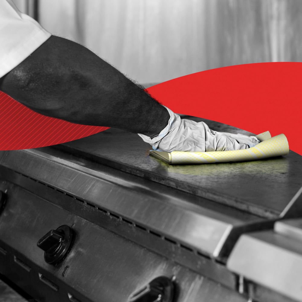 En person tørker av en kommersiell kjøkkenkomfyr med hansker med en gul WypAll® X80 Critical Clean - Interfold (41 x 33 cm) fra Kimberly-Clark GmbH; i bakgrunnen vises et rødt grafisk element.