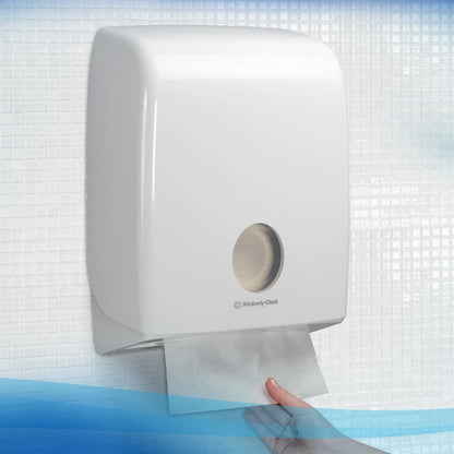 Una mano saca una toalla plegada Scott® Control™ de Kimberly-Clark GmbH de un dispensador de pared blanco con ventana redonda frente a un fondo de azulejos blanco-azul. Empaquetado: 15 clips x 240 hojas por caja (15 paquetes).