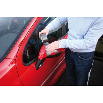 Eine Person in einem hellblauen Pullover verwendet das Scotch® Extremium™ No Residue Hochleistungs-Gewebeklebeband (18,2 m x 48 mm) von 3M, um den beschädigten Seitenspiegel eines roten Autos zu befestigen, während sie an der Fahrertür steht. Das graue Band ist effektiv und hinterlässt keine Rückstände.