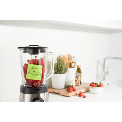 En mikser med jordbær står på en hvit kjøkkenbenk. En Post-it® Super Sticky Note (76 x 76 mm, 90 ark, 100 % PEFC) fra 3M Deutschland GmbH er festet til beholderen. I nærheten er redskaper, potteplanter, olivenolje og flere jordbær på et skjærebrett.