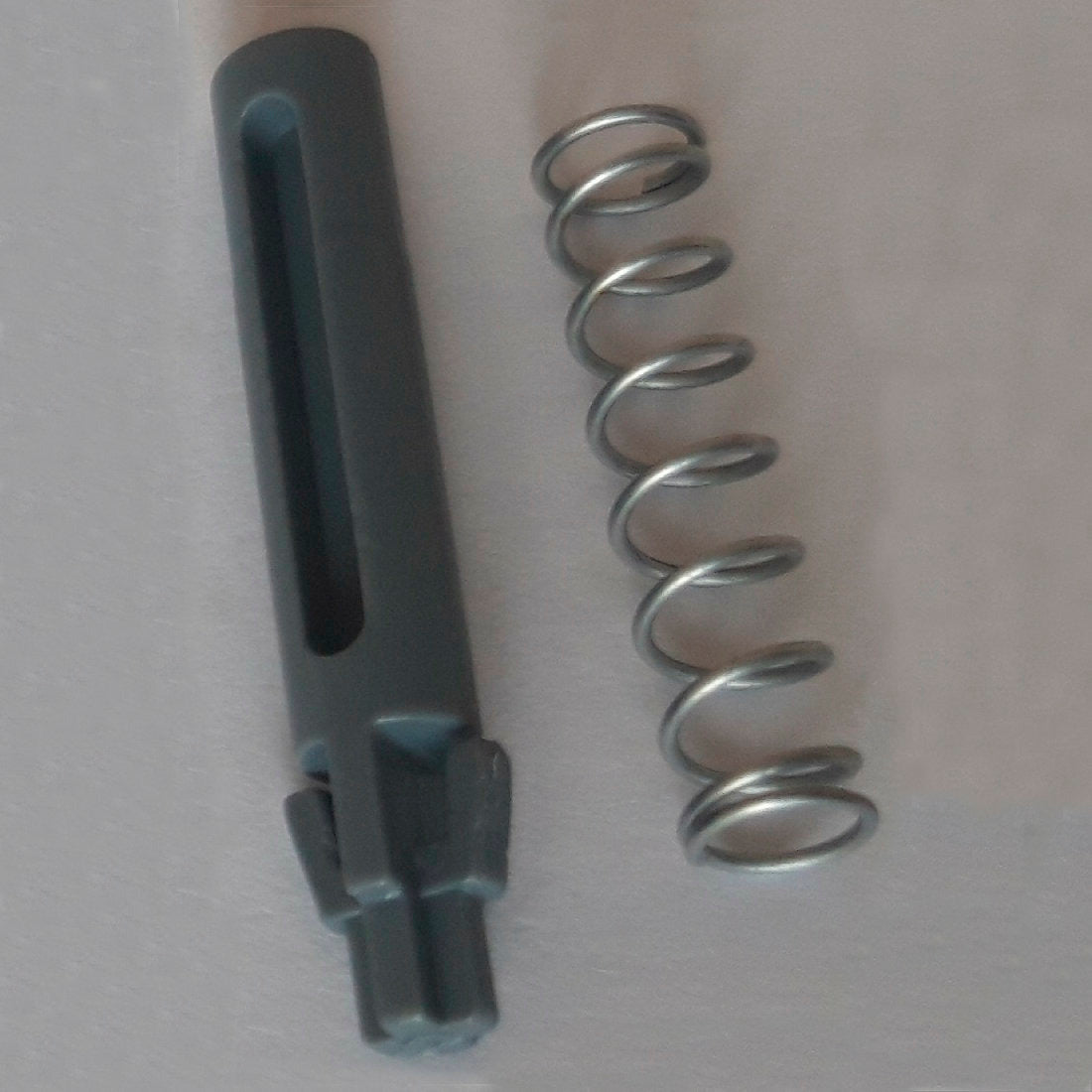 Un resorte de metal y un pasador de puerta cilíndrico de repuesto de color gris oscuro con ranuras y muescas, parte del pasador y resorte de puerta DI Protect (1 pieza) de Diversey Deutschland GmbH & Co. OHG, están representados sobre una superficie clara y muestran los materiales robustos para el carrito DI Protect.