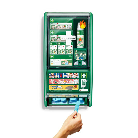Eine grüne Cederroth erste hilfe & verbrennungsstation von Orkla Wound Care AB ist an einer weißen Wand montiert, bestückt mit Verbänden, Verbrennungsgel, Augenspülung und mehr. Eine Hand zieht eine blaue Klebebinde aus dem unteren Spender des 1er-Kartons.