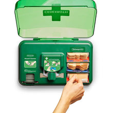 Ein grüner, wandmontierter Orkla Wound Care AB Auslaufartikel: Cederroth Wound Care Dispenser (20,3x30,6x15,5 cm) mit offenem Deckel zeigt Wundreiniger und Verbandsmaterial, während eine Hand einen Verband aus dem Pflasterspender Folienbeutel-Set zieht.