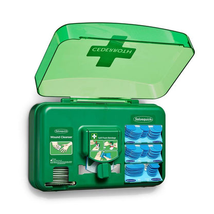 Ein blauer Wundversorgungsspender von Orkla Wound Care AB ist geöffnet und gibt den Blick frei auf Wundreinigungspakete, Pflaster und eine Rolle weicher Schaumstoffbinden, die in einzelnen Fächern angeordnet sind.