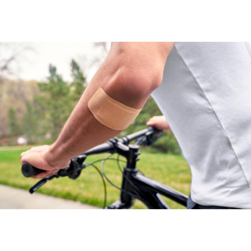 Una persona que lleva una camisa blanca y monta en bicicleta al aire libre tiene un apósito Nexcare™ Ultra Stretch MAXI Comfort Flexible (50 mm x 101 mm) de 3M Deutschland GmbH en el brazo desnudo; al fondo se ven césped verde y árboles.