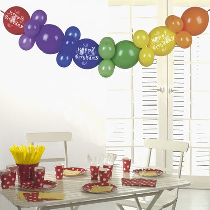 Ein weißer Tisch mit rot gepunktetem Partygeschirr, gelben Utensilien und Bechern wird von der PAPSTAR Ballongirlande 2 m "Happy Birthday" der PAPSTAR GmbH gekrönt - eine festliche Partydekoration Geburtstag, die im Sonnenlicht durch weiße Fensterläden leuchtet.