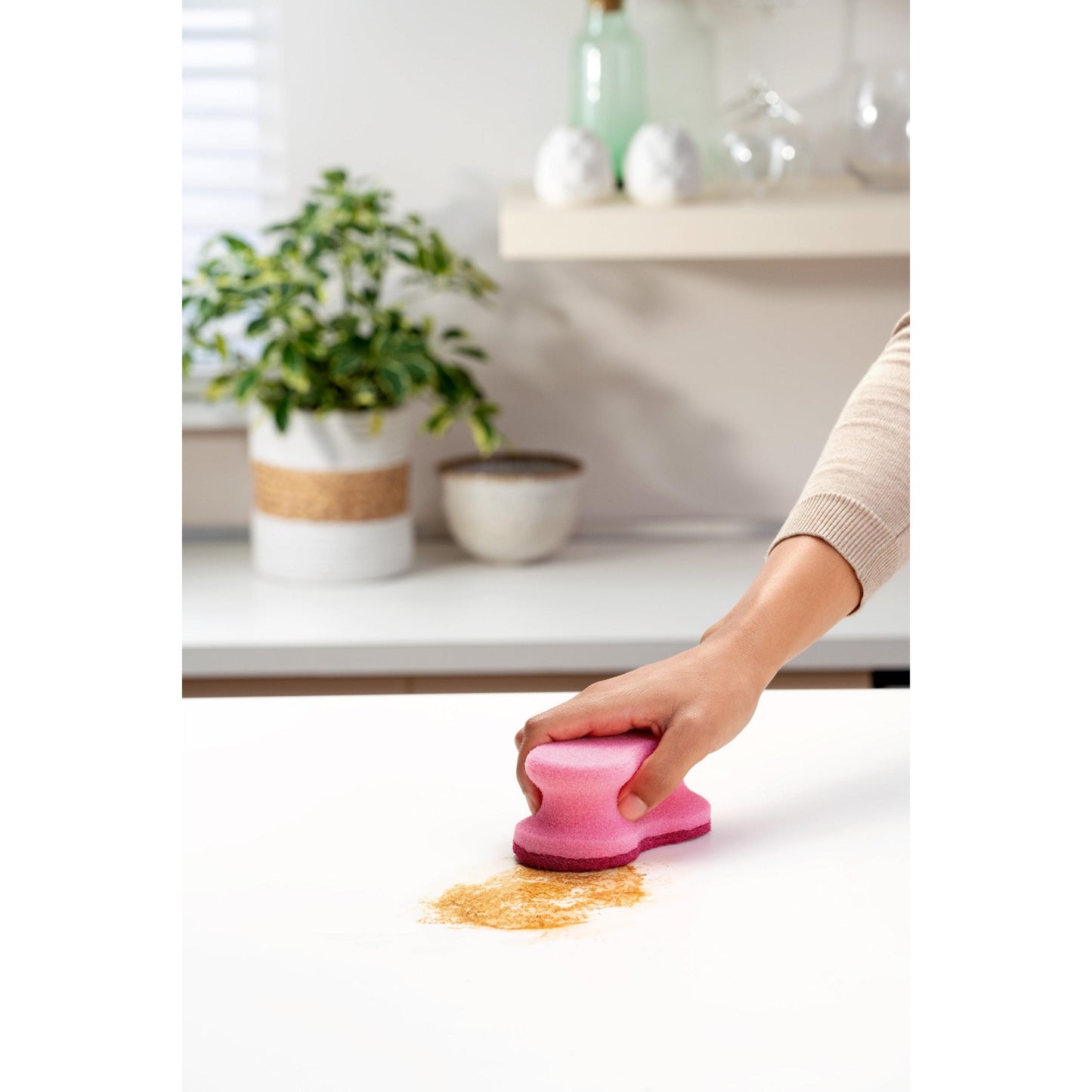 Eine Hand benutzt einen rosafarbenen Scotch-Brite® Delicate Color Comfort nicht kratzender Reinigungsschwamm (2 Stück, 3M Deutschland GmbH), um braunes Pulver von einer weißen Arbeitsplatte zu reinigen - perfekt für empfindliche Oberflächen. Pflanze und Schalen im Hintergrund.