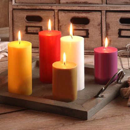 Five burning PAPSTAR pillar candles (Ø 70 mm, 150 mm, 100% stearin, fully colored) from PAPSTAR GmbH in the colors yellow, red, white, orange, and pink are arranged on a wooden tray with handles on a wooden table.