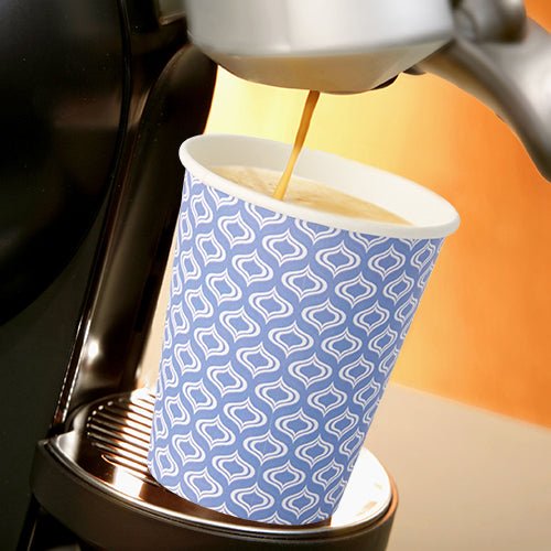 A Starpak 20 “To Go” cup made of recyclable cardboard (0.2 l, Ø 8–9.1 cm) with a blue-and-white pattern sits beneath the spout of a coffee machine that’s pouring fresh coffee. The scene is brightly lit.