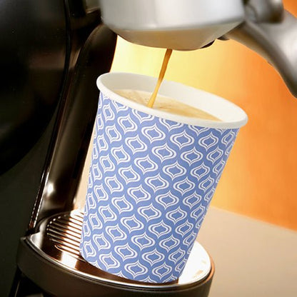 A Starpak 20 “To Go” cup made of recyclable cardboard (0.2 l, Ø 8–9.1 cm) with a blue-and-white pattern sits beneath the spout of a coffee machine that’s pouring fresh coffee. The scene is brightly lit.