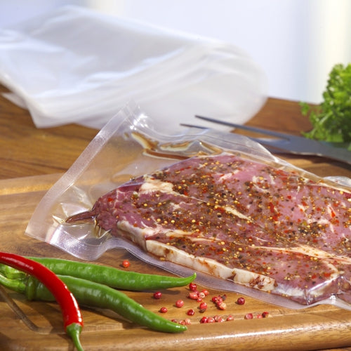 Ein Starpak 100 Siegelrandbeutel, PA/PE transparent von Starpak hält mariniertes rohes Fleisch auf einem Holzbrett mit Chilis und Grünzeug - ideal zum Vakuumieren von Lebensmitteln mit einem Kammervakuumierer.