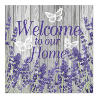 Sign with purple text reading "Welcome to our Home" on a gray wooden background, decorated with lavender-colored flowers and three white butterflies, inspired by PAPSTAR 20 napkins, 33x33 cm, 3-ply, from PAPSTAR GmbH.