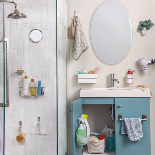 Ein Badezimmer mit einer Glasdusche auf der linken Seite, geordneten Regalen, einem Waschbecken mit blauem Schrank darunter, in dem Reinigungsmittel aufbewahrt werden, einem ovalen Spiegel darüber und einem 3M Command™ Großer Doppelhaken von 3M Deutschland GmbH, der ein Handtuch am Schrankgriff hält.