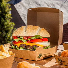 A cheeseburger with lettuce, tomato, onion, and cheddar cheese lies outdoors in a PAPSTAR GmbH burger box made of cardboard (9x18.5x18.5 cm, "100% Fair", 25 pieces). French fries and other side dishes are served separately next to it.