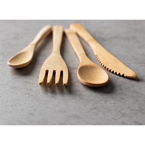 Cuatro utensilios de madera - una cuchara, un tenedor, otra cuchara y un cuchillo de bambú PAPSTAR "pure" de 16 cm - están dispuestos sobre una superficie de piedra gris. Los cuchillos desechables ecológicos de PAPSTAR GmbH son de color marrón claro y tienen una superficie lisa y pulida.