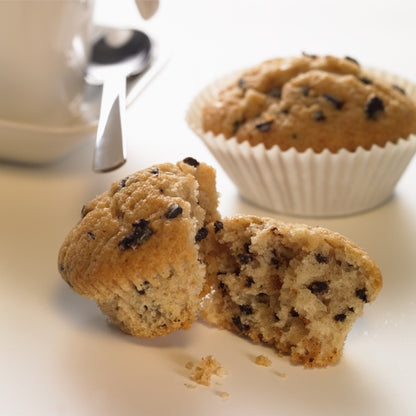 Im Vordergrund ist ein halbierter Chocolate Chip Muffin zu sehen, dahinter ein ganzer Muffin in einem PAPSTAR 1000 Backförmchen rund der PAPSTAR GmbH. Im Hintergrund sind eine weiße Tasse mit Untertasse und Löffel angeschnitten.