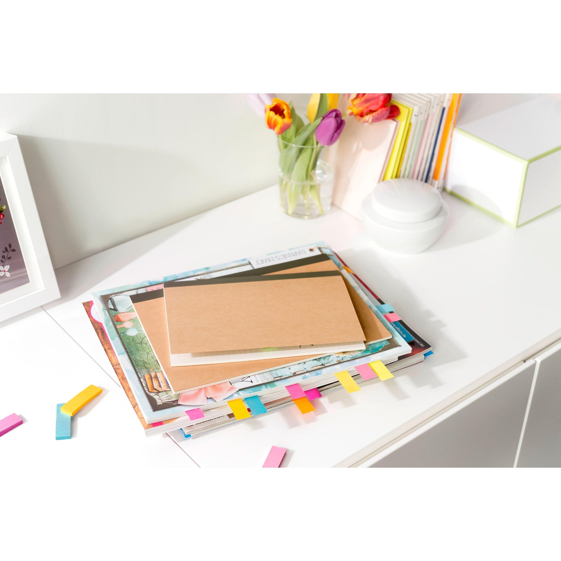 Ein Stapel Zeitschriften und ein Notizbuch mit farbenfrohen Post-it® Page Markern, Poptimistic Collection von 3M Deutschland GmbH (15 mm x 50 mm, 4 Blöcke/Packung), liegen auf einem weißen Schreibtisch neben Tulpen und aufrecht stehenden Büchern und schaffen ein geordnetes, helles Ambiente.