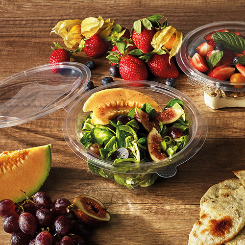 Auf einem Holztisch steht eine runde Starpak 50 Feinkost- und Salatschalen mit Klappdeckel (R-PET glasklar) mit Salat aus Melone, Feigen und Blattgemüse. Daneben stehen Erdbeeren, Weintrauben, Melonenscheiben, Kirschtomaten, Fladenbrot und eine weitere Schüssel mit Tomaten und Basilikum.