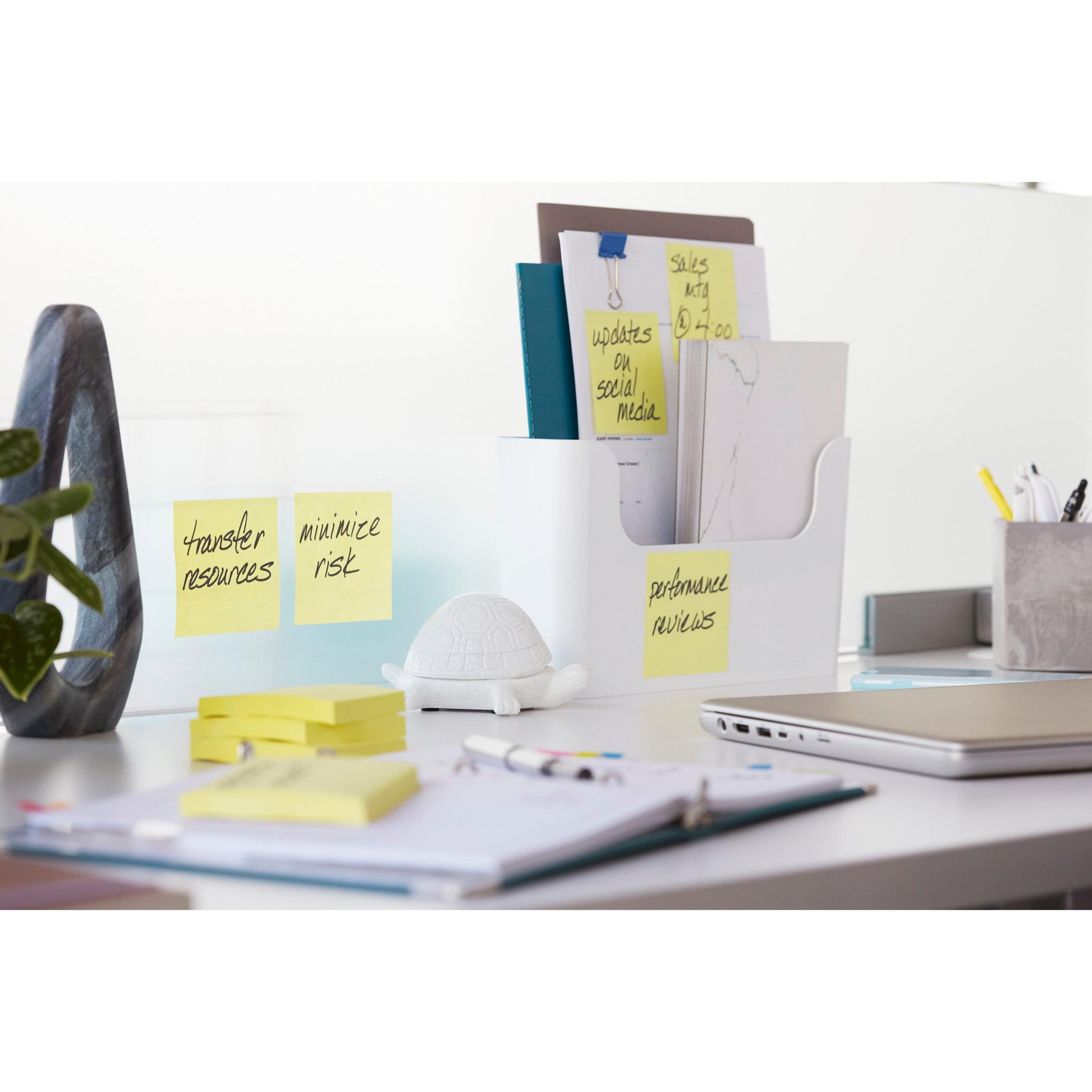 Ein weißer Schreibtisch mit dem Post-it® Z-Notes Dispenser PRO der 3M Deutschland GmbH in Schwarz mit gelben Post-it® Super Sticky Z-Notes (76x76 mm), dazu Mappen, Büromaterial, ein Notebook, ein Laptop, eine Schildkrötenfigur und Haftnotizen an der Wand, auf denen Aufgaben aufgelistet sind.