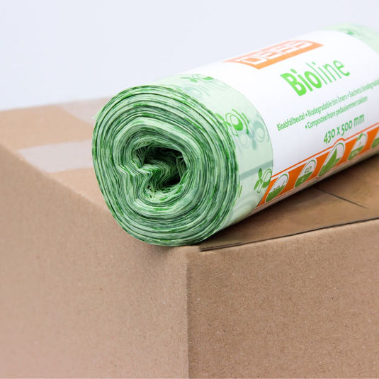 A roll of green compostable waste bags is placed on a cardboard box. The label on the roll is partially visible and bears the print "EMIL DEISS KG (GmbH + Co.)". These 100% compostable bags have a floral design and measure 430 x 500 mm.