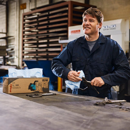 A man in a dark blue overall wipes a tool with a WypAll® X80 PowerClean (BRAG™ Box / Steel Blue) from Kimberly-Clark GmbH at an industrial workbench, with a box of disposable towels, tools, and shelves in the background.