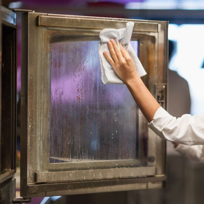 A person in a white shirt uses the Tork 570137 Extra Strong Industrial Cleaning Cloths W1 W2 W3 1-ply from TORK – Essity Professional Hygiene Germany GmbH to clean an oven glass door with a cloth. The oven is open, showing steam and grease stains, while the background is blurred with a hint of purple and other colors.
