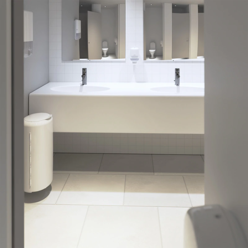The modern public restroom features an elegant white vanity top with two sinks and chrome fixtures. On the left, there is a white trash can, while mirrors hang above the tiled floor. Tork Xpress® 552000 soap dispensers by Essity Professional Hygiene Germany GmbH are mounted on the wall, and Multifold hand towels enhance the comfort of visitors.