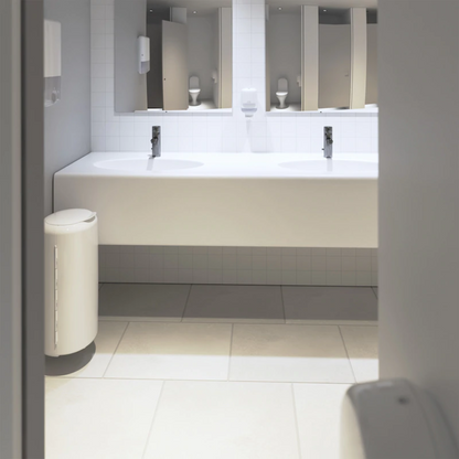 The modern public restroom features an elegant white vanity top with two sinks and chrome fixtures. On the left, there is a white trash can, while mirrors hang above the tiled floor. Tork Xpress® 552000 soap dispensers by Essity Professional Hygiene Germany GmbH are mounted on the wall, and Multifold hand towels enhance the comfort of visitors.