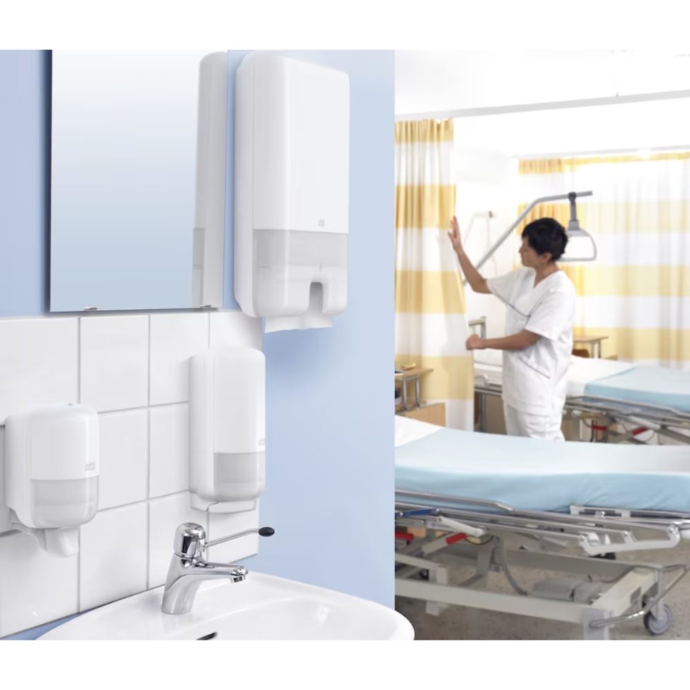 In a hospital room, a doctor in a white uniform stands next to a bed. In the foreground, there is a sink equipped with a Tork Xpress® 552000 dispenser for Multifold Hand Towels H2 by TORK – Essity Professional Hygiene Germany GmbH. The room is equipped with medical devices and yellow striped curtains.