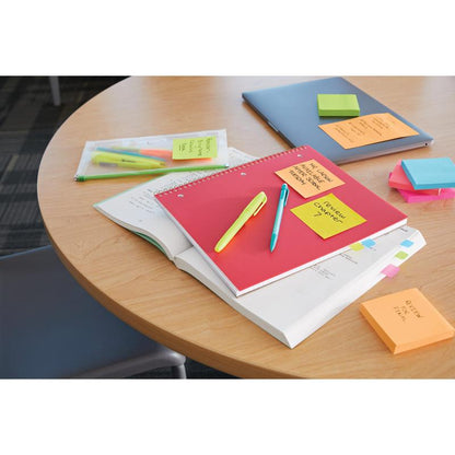 A round wooden table with a closed laptop, notebooks, pens in a plastic bag, highlighters, reminder notes, and a stack of 3M Deutschland GmbH Post-it® Super Sticky Notes Cube, Yellow (76 mm x 76 mm). A chair is partially visible.