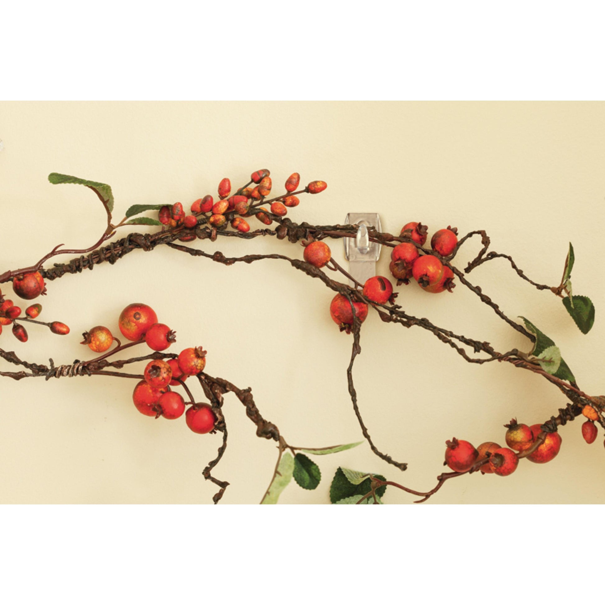 Rote Beeren und grüne Blätter an braunen, gedrehten Zweigen werden mit einem 3M Command™ Mini-Haken, transparent, mit transparenten Streifen (17006CLRFGN), der in der Mitte des Bildes zu sehen ist, an eine hellgelbe Wand gehängt.