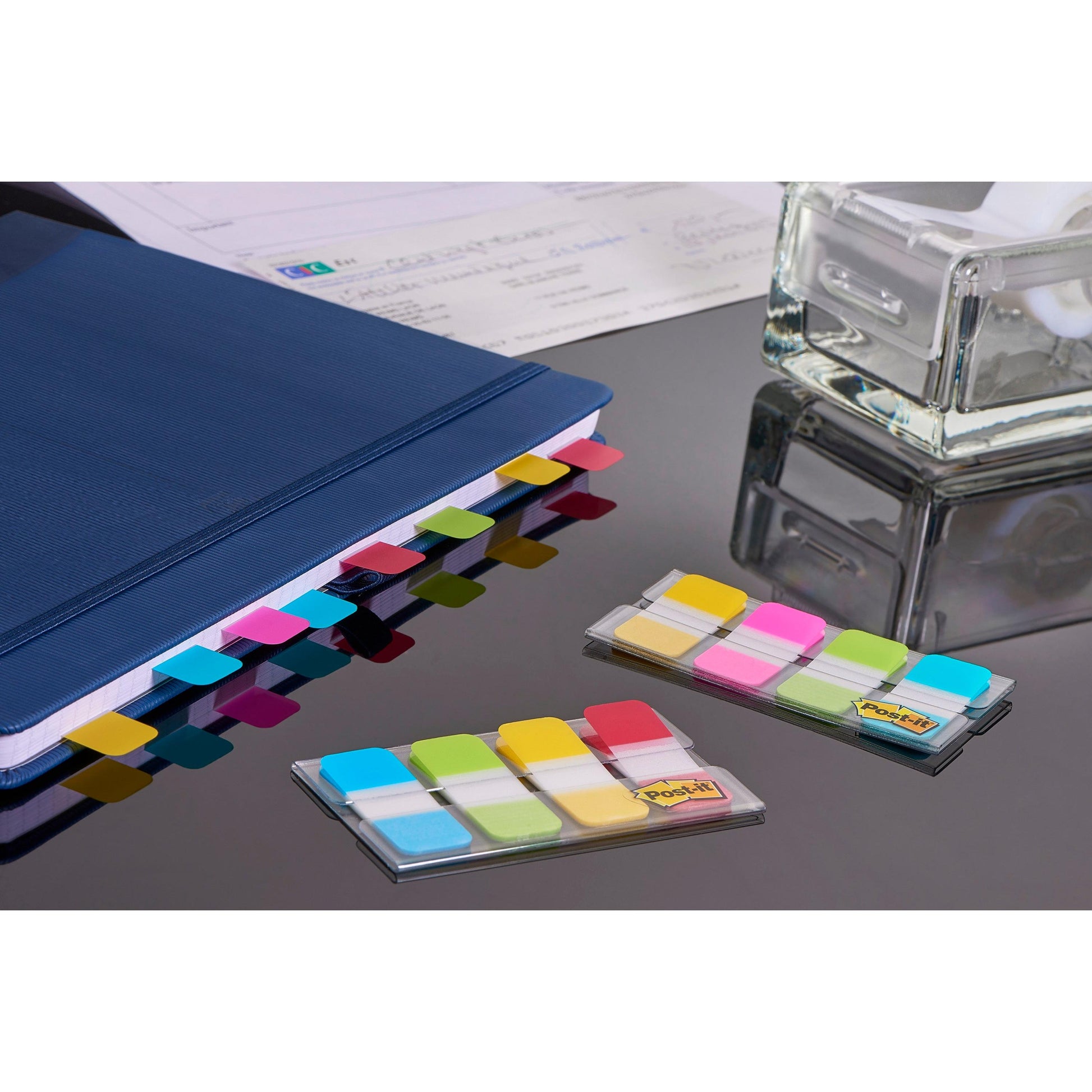 Ein blaues Notizbuch mit Post-it® Index Strong Ablagetabs (Mini, Blau, Violett, Rosa, Gelb; 15,8 mm x 38,1 mm) von 3M Deutschland GmbH liegt auf einem Glastisch zusammen mit Papierblättern, einem Klebebandabroller und zusätzlichen Packungen von Ablagetabs.