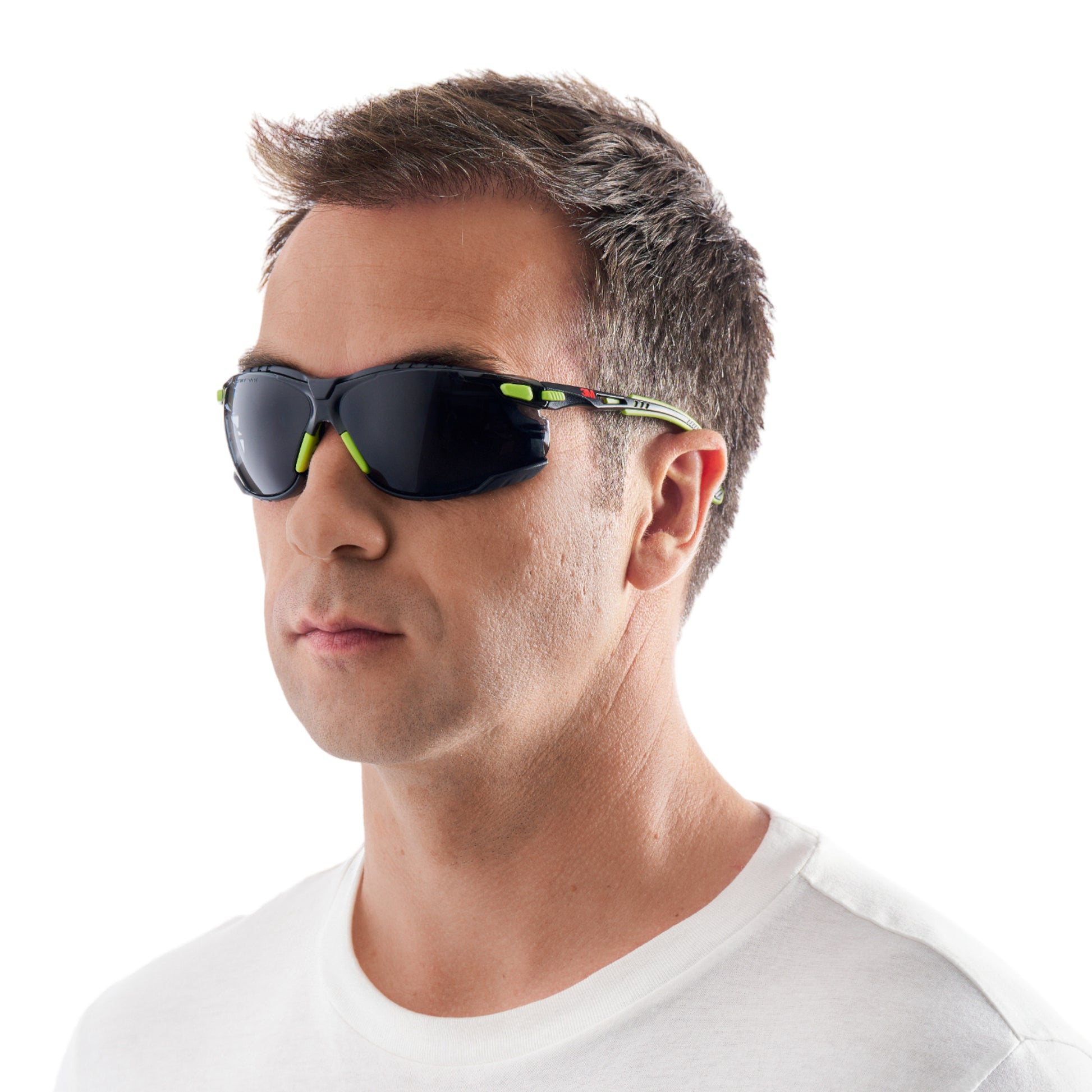 Ein Mann mit kurzen braunen Haaren trägt eine 3M™ Solus™ Schutzbrille 1000 Sonnenbrille in grün/schwarz mit grauen Gläsern von 3M Deutschland GmbH, gepaart mit einem schlichten weißen T-Shirt, mit Blickrichtung leicht nach links vor einem weißen Hintergrund.
